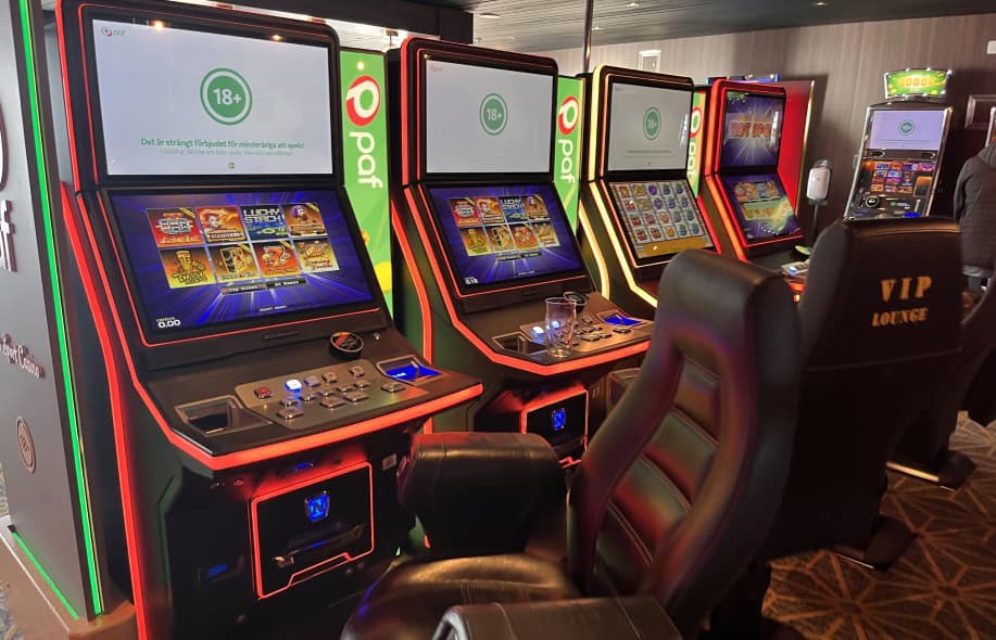 Kuvassa näkyy rahapeliautomaatteja rivisä. Penkissä lukee "VIP Lounge". Aiempi pelaaja on jättänyt lasituopin yhden automaatin päälle. Pelien kyljessä Paf logo. Taustalla näkyy käsidesinfiointipiste.