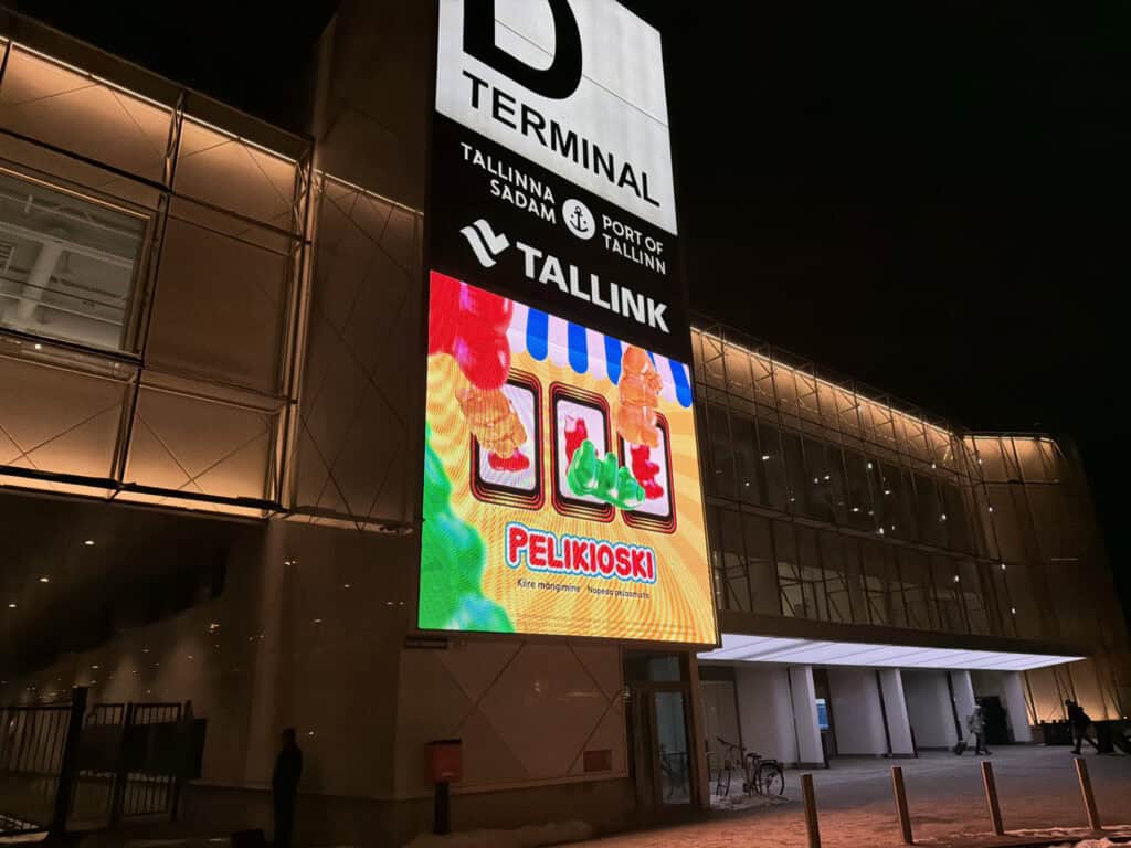Tallinnan sataman D-terminaalin rakennus ulkoapäin kuvattuna ilta-aikaan. Seinässä kiinni iso mainoskyltti missä Pelikioski nettikasinon hedelmäpeli näyttää sylkevän ulos nallekarkkeja. Mainoksessa tulee tunne R-kioskista sen värityksen takia.