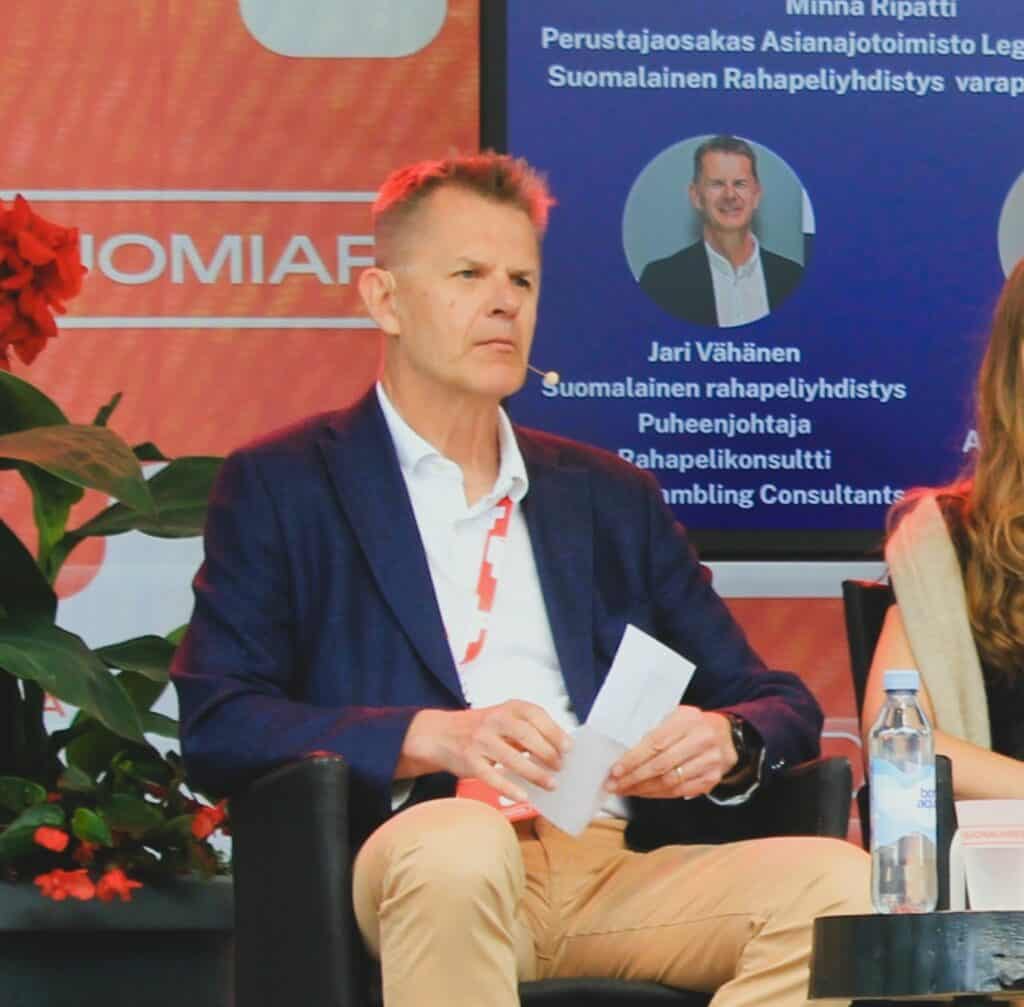 Jari Vähänen panelistossa Suomi Areena 2026 tapahtumassa. Taululta näkyy että samassa paneelissa Minna Ripatti. Vieressä toinen nainen. Vähäsen titteli on Suomalaisen rahapeliyhdistyksen puheenjohtaja ja rahapelikonsultti Finnish Gabmling Consultants.