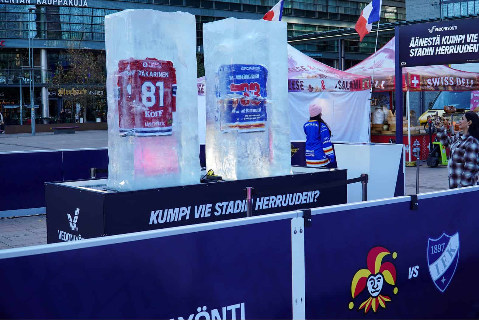 Veikkauksen mainospisteellä sai äänestää Helsingin jääkiekkoderbyn suosikistaan. Jokerit vs. HIFK. Suuret Veikkaus-mainoskyltit. Narinkkatorilla tiistaina 6. lokakuuta 2026. Symbolisoi rahapelien ajankohtaisuutta – Suomessa siirrytään valtion kontrolloimista peleistä valtion lisensoimiin peleihin. Taustalla juustokoju ja Simonkentän rakennuksia.