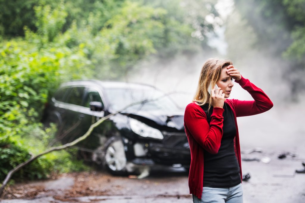 A young woman on the phone in front of her damaged car with a tree on top of it.