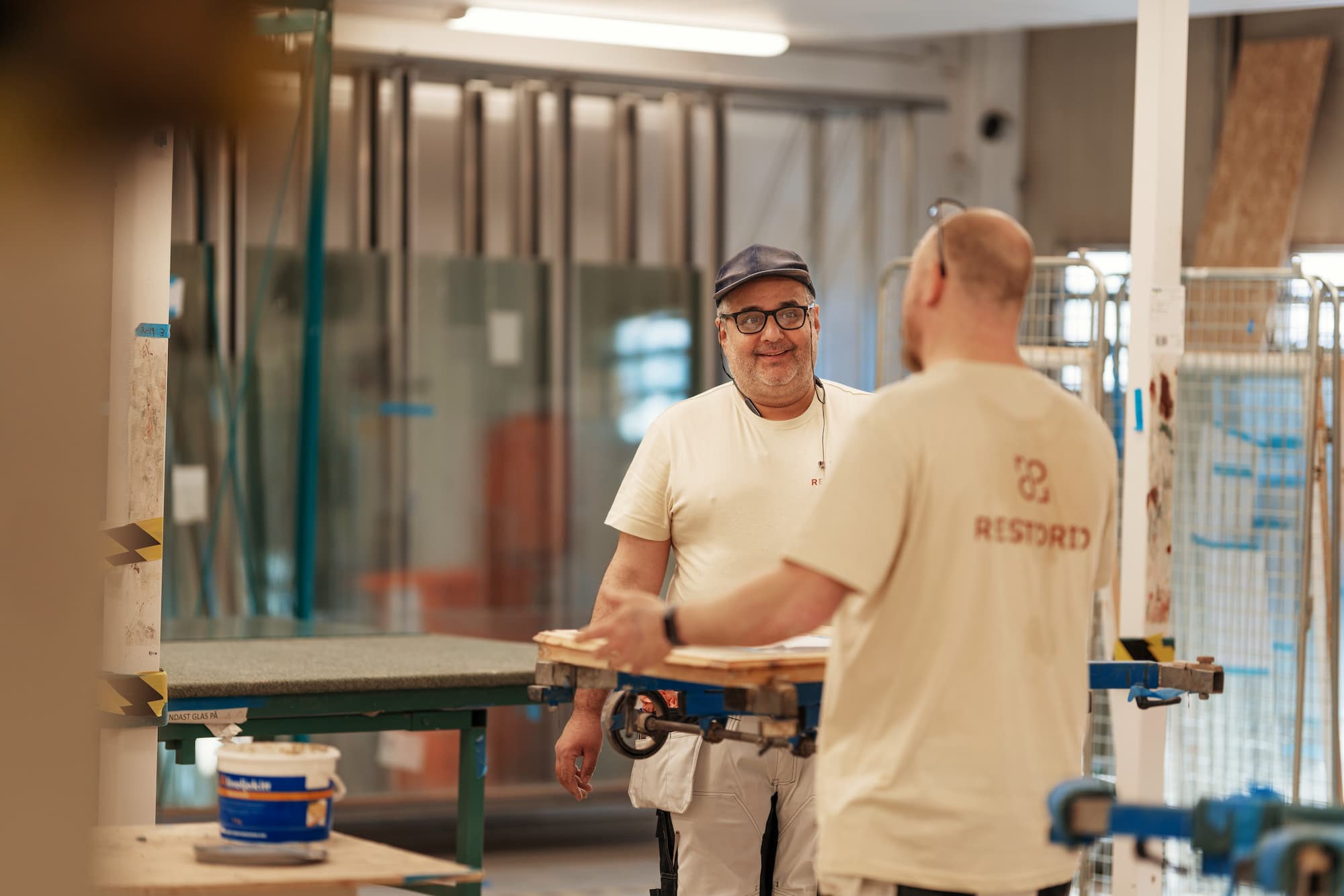 Två medarbetare från Restorio samarbetar i glasmästeriet, en av dem bär skyddsglasögon och ler mot kameran.