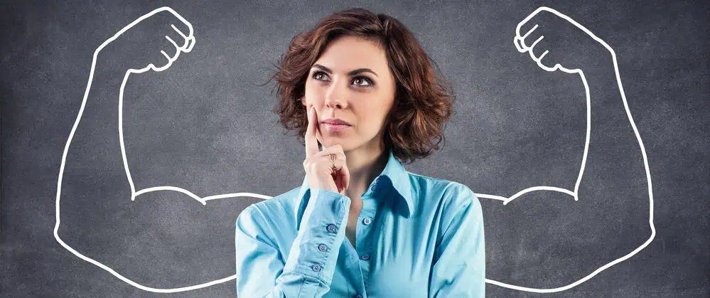 Strong woman Concerned looking woman standing in front of a blackboard that has arms with huge flexing biceps drawn on it, so that it looks like they are the woman's arms