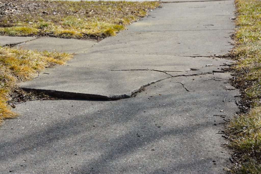 Cracked and uneven sidewalk with grass on both sides, in need of repair.