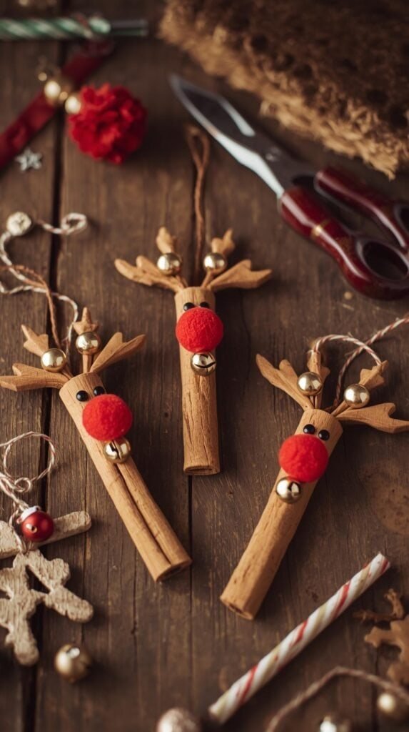 Three cinnamon stick reindeer ornaments displayed on a rustic wooden table with craft supplies beside them. Red pom-pom noses and jingle bells add festive touches.