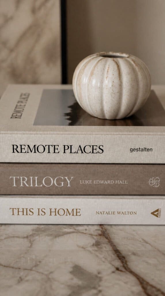 Three coffee table books stacked horizontally on marble surface with a small ceramic object placed on top, warm neutral cover tones, editorial flat lay photography