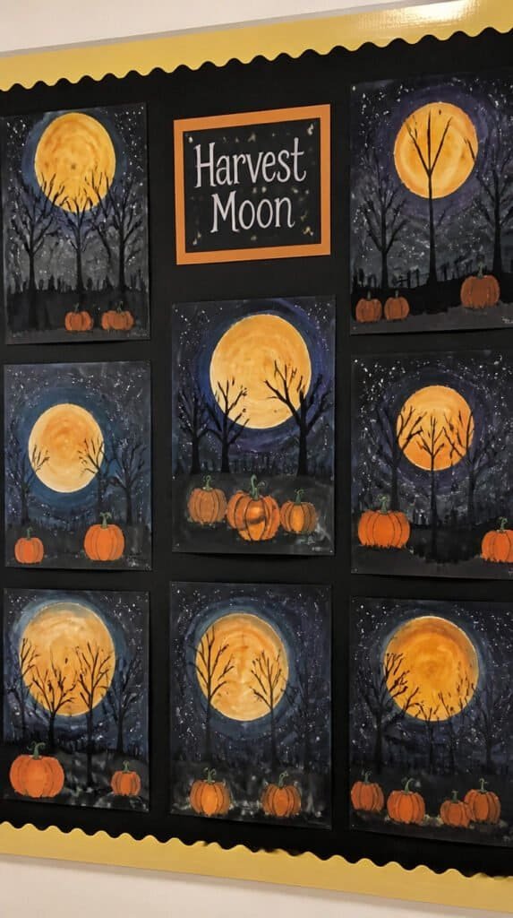 Harvest moon watercolor art on dark blue paper showing glowing gold moon over silhouette of bare trees and pumpkins