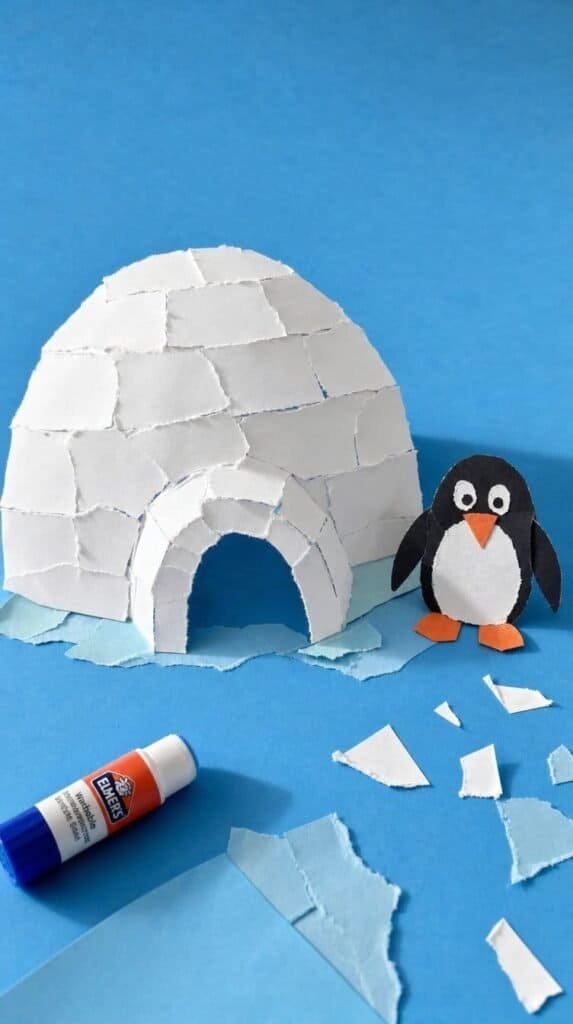 Torn white paper igloo collage on blue paper with a small paper penguin