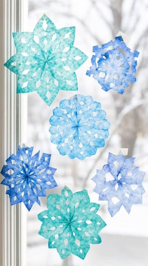 Blue and teal dyed coffee filter snowflakes taped to a sunny window