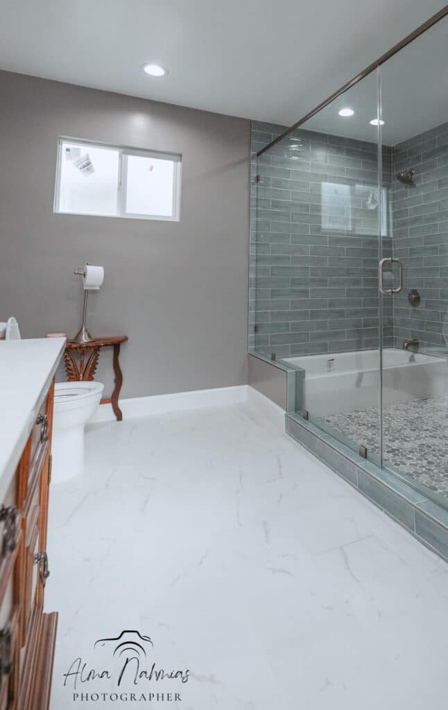 Modern bathroom with glass shower, white tub, and gray tiled walls.