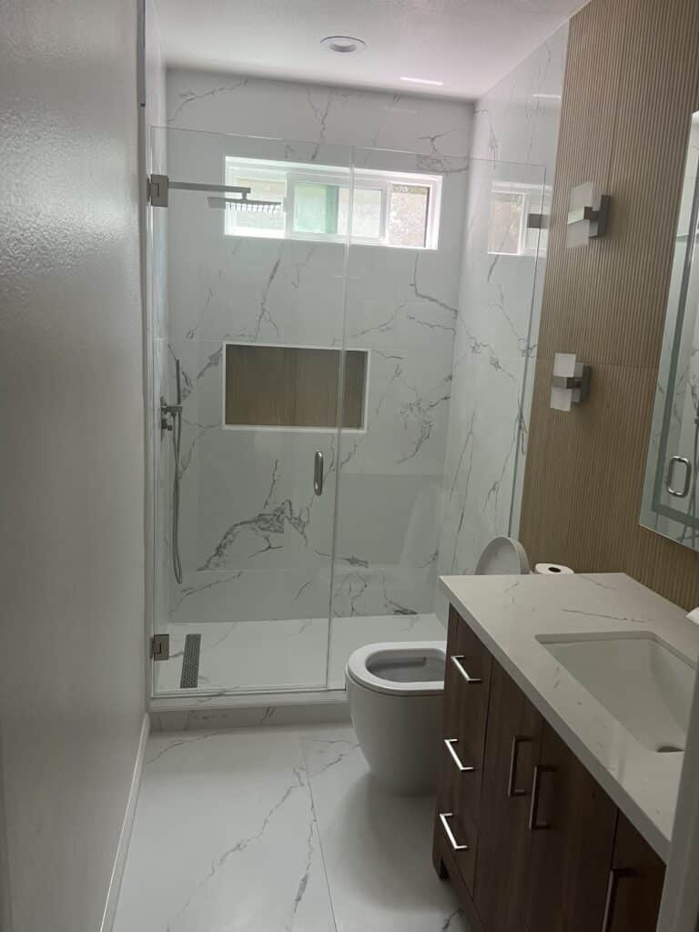 Modern bathroom with white marble tiles, glass shower, and wooden vanity.