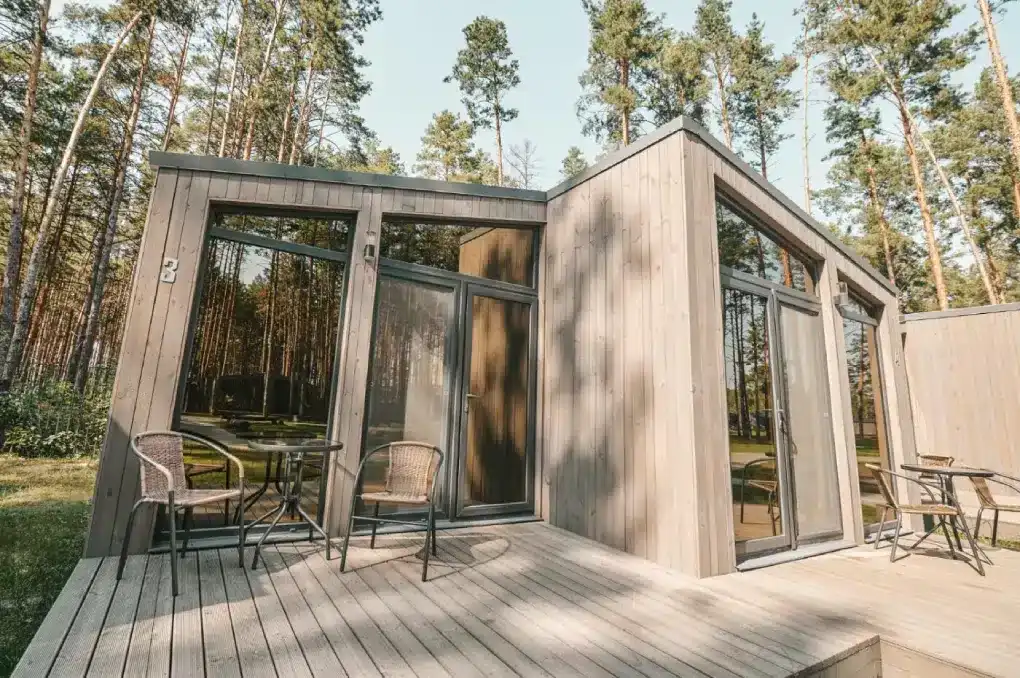 Modern wooden cabin with large windows and outdoor seating in a forest.