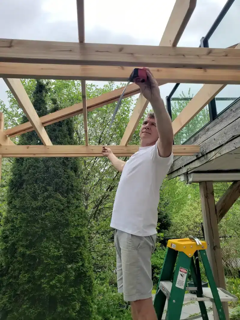 Man in white shirt measuring wooden beams for an outdoor structure.