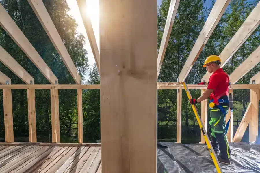 Construction worker in a red shirt and yellow hard hat measuring a wooden beam.