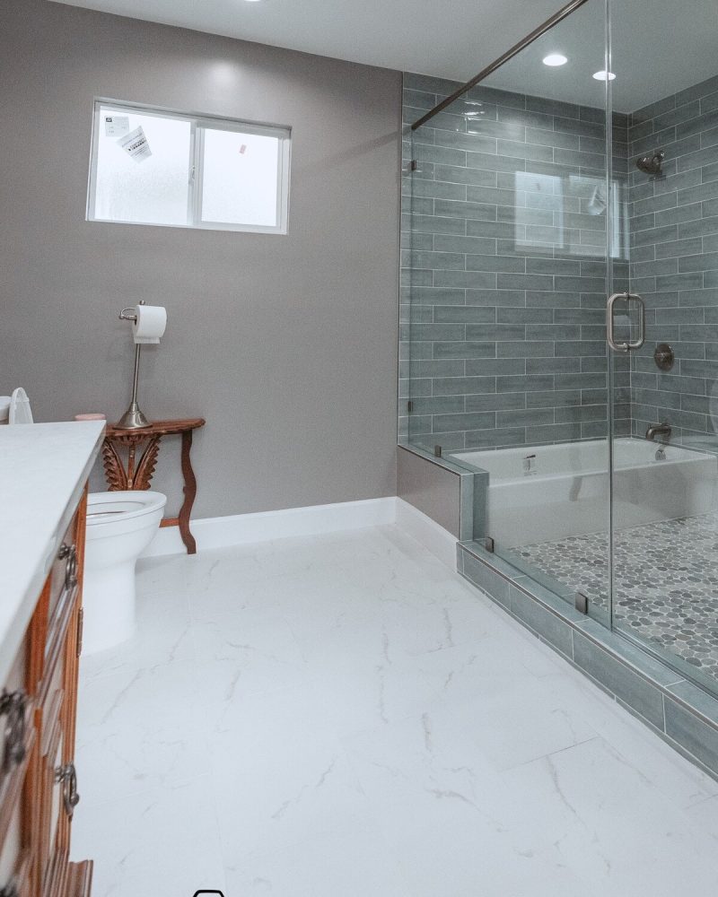 Modern bathroom with glass shower, white tub, and gray tiled walls.