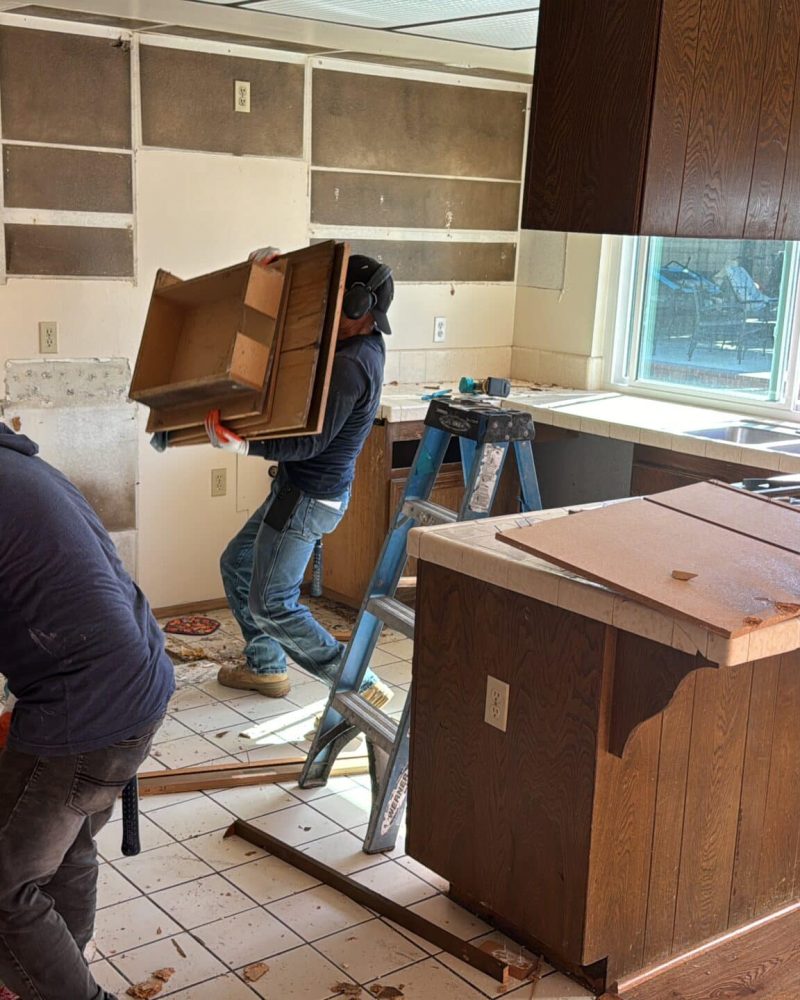 Two workers remove cabinets from a kitchen undergoing demolition.