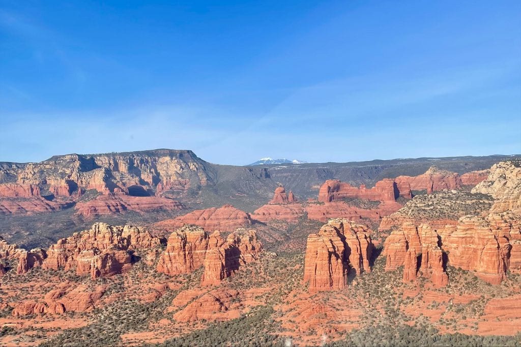 This image features a breathtaking view of a desert landscape, showcasing the distinct red rock formations of Sedona under a clear blue sky. The rugged terrain is marked by numerous steep cliffs and deep valleys, with a backdrop of distant mountains partially covered in snow, illustrating a vivid contrast between the arid foreground and the icy peaks.