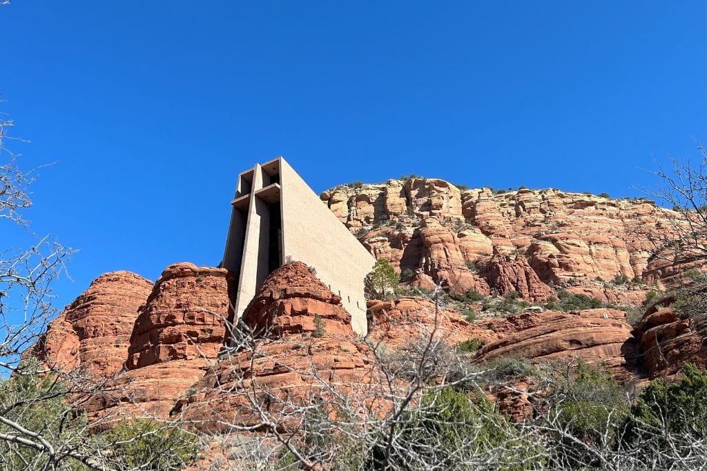 The image features the Chapel of the Holy Cross, an architectural marvel, built into the red rock formations of Sedona, Arizona. The modernistic structure, characterized by a prominent cross and angular designs, contrasts starkly against the natural, rugged red cliffs under a clear blue sky.