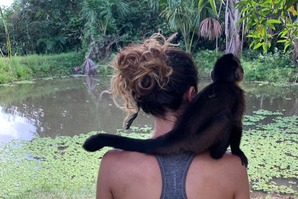 Giorgy with curly hair stands with their back to the camera, a black Howler monkey perched on their shoulder, both overlooking a tranquil waterbody covered with green foliage. 