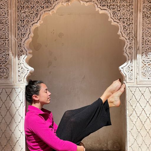 The Seven Saints of Marrakech (Local Insight of Marrakesh) Giorgy, the woman in a bright pink jacket and black pants is performing a yoga pose in an intricately carved stone alcove. The alcove features detailed geometric patterns, providing an ornate backdrop to the serene scene.
