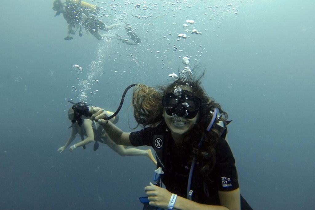 A group of scuba divers is captured exploring the underwater world; one diver with a beaming smile is in the foreground, her hair floating around as bubbles ascend around her. The backdrop is a dimly lit, expansive ocean, hinting at the vastness and mystery of the deep blue.