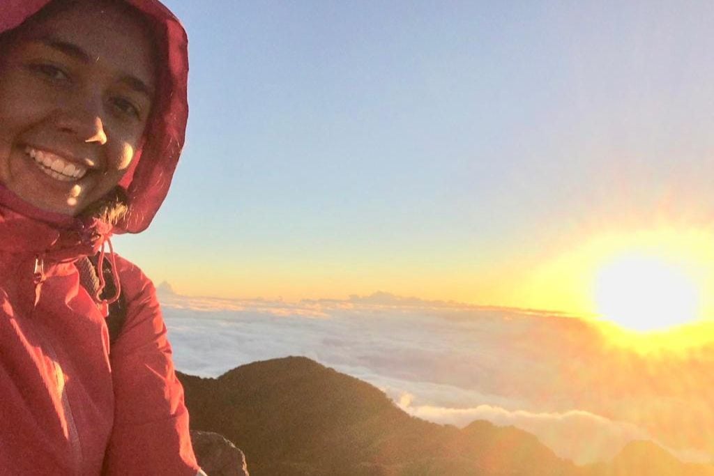 The 2026 Spiritual Guide to Boquete Panama A selfie of a smiling person wearing a red hooded jacket with the sun rising behind them, casting a warm glow over a sea of clouds and distant mountains on the Volcan Baru. Spiritual Guide to Boquete Panama.