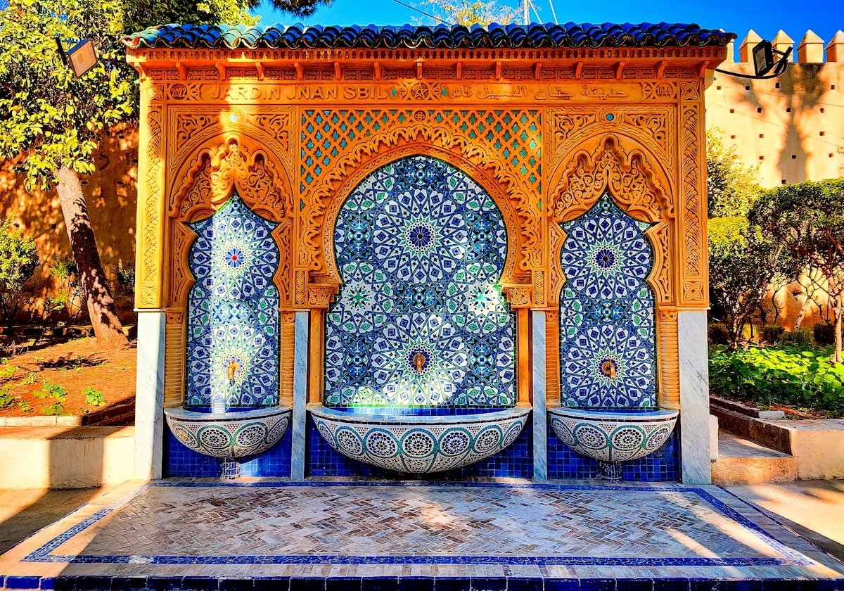 An ornate water fountain in Jardin Jnan Sbil of Fes, Morocco, with triple arches adorned in vibrant blue, green, and orange zellige tilework, sits under a canopy of trees against a sunlit terracotta wall. The fountain's reflection on the wet tiled floor adds to the tranquil ambiance of the park.