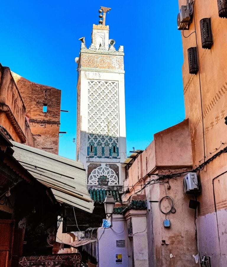 A tall white minaret adorned with intricate green and blue mosaic patterns towers above the terracotta-colored buildings of the medina, under a clear blue sky. Electrical wires, air conditioning units, and satellite dishes accent the textured walls of the surrounding structures. Discover the rich culture of Fez Morocco.