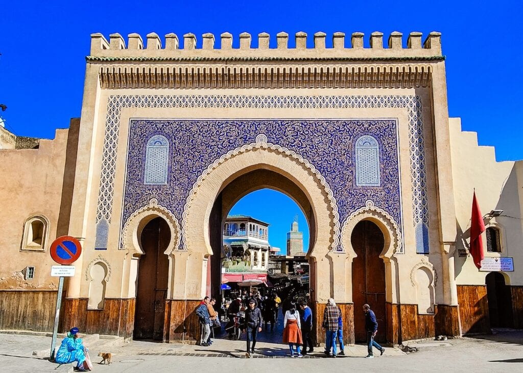 The historic Bab Bou Jeloud gate in Fes features a grand horseshoe archway framed by intricate blue tilework, with a bustling street scene visible through the gate. People walk by, some pausing to converse, under a vibrant blue sky, with Moroccan architecture and the city's distinctive terracotta buildings in the background. Picture of Fez Morocco rich culture. 