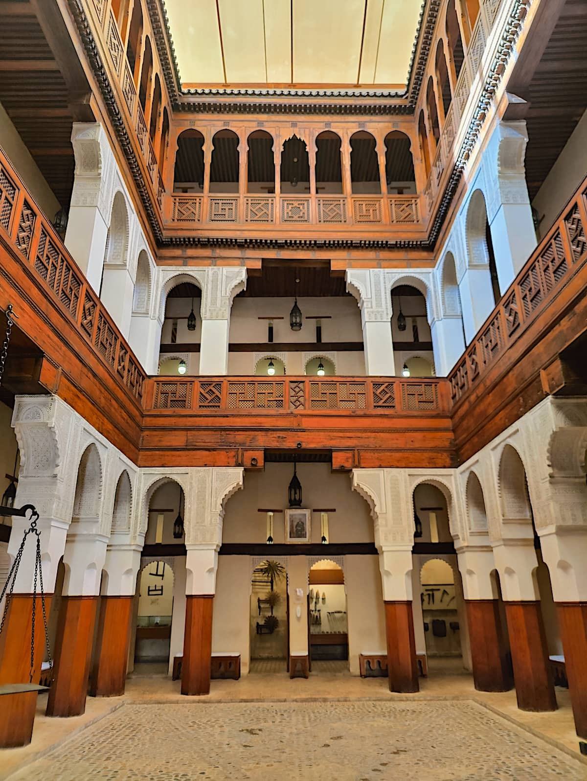 Interior view of a museum in Fes, Morocco, displaying traditional Moroccan architecture with a three-tiered wooden gallery, ornate carved stucco, and intricately patterned dark wood balconies. Elegant arches lead to serene inner rooms, beneath a vast wooden ceiling, creating a sense of historical grandeur.