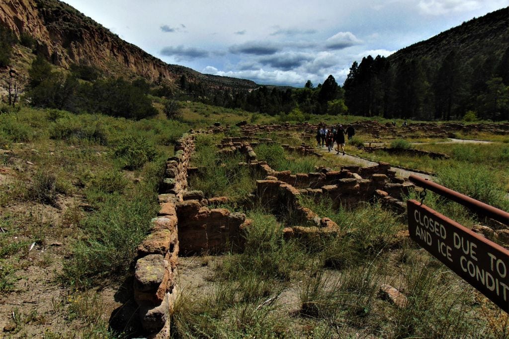 Your Guide to Santa Fe Spiritual Vortex in 2026 Tourists walking by an ancient archaeological site with ruins of stone structures, a sign reads 'CLOSED DUE TO WET AND ICE CONDITIONS', emphasizing the historical context of the location. used for the article a guide to Santa Fe spiritual Vortex.