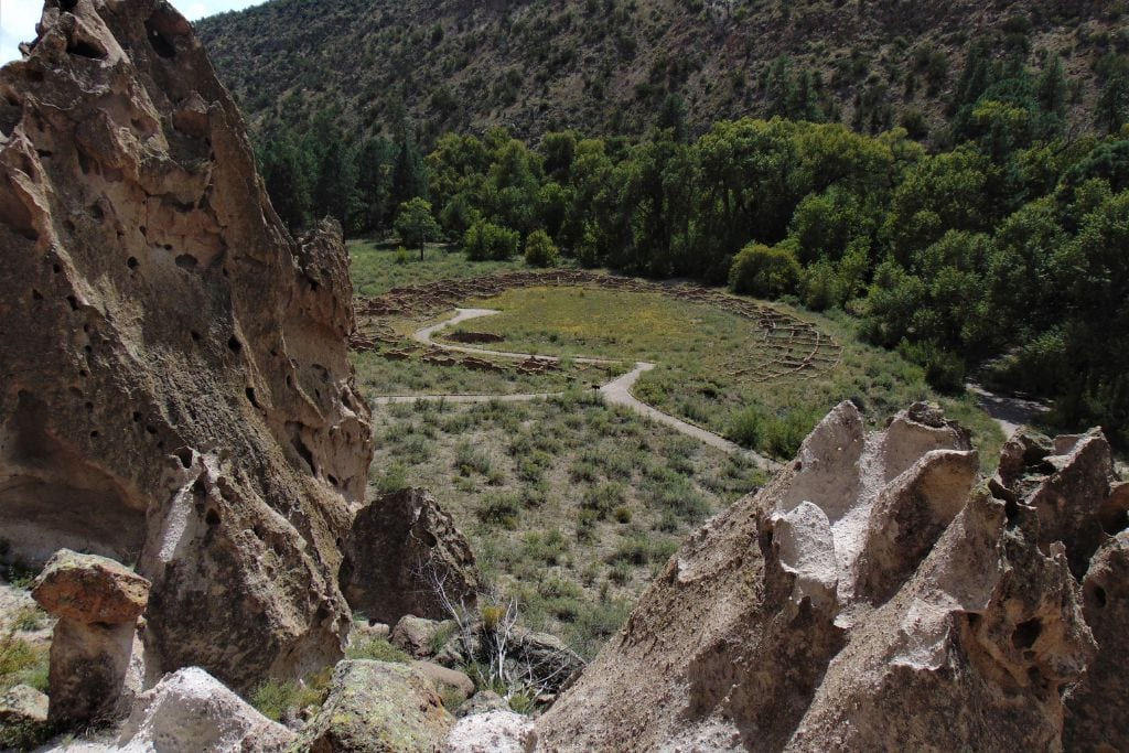 Rugged rock formations frame a serene view of a winding path through greenery, leading to a circular structure in the distance, evoking the essence of a spiritual quest in Santa Fe's natural landscape. Santa Fe Spiritual Vortex in 2025