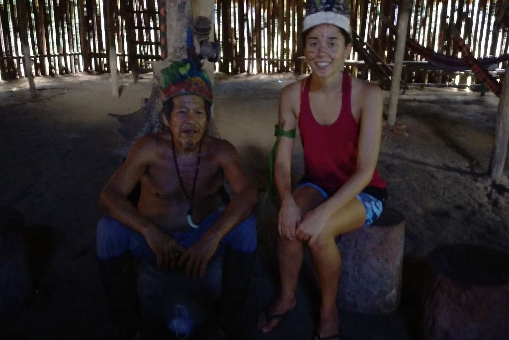 Two individuals are sitting on wooden stumps inside a dimly-lit traditional hut with a bamboo wall structure. The person on the left wears a colorful headpiece and a necklace, suggestive of indigenous attire, while the person on the right is smiling broadly, dressed in casual clothing. The ambiance suggests a cultural interaction or a visit to a local community.