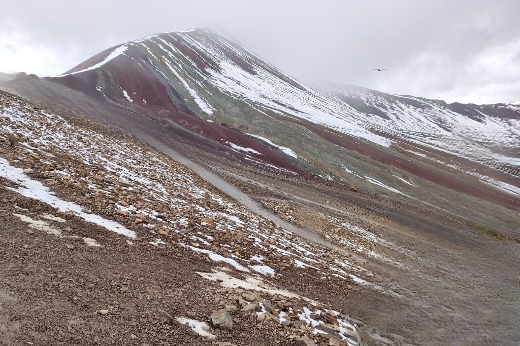 Rainbow Mountain Peru: Your Ultimate Spiritual Guide Dynamic view of Rainbow Mountain's slopes under a misty sky, showcasing layers of snow-dusted ground, leading up to the mountain's vibrant, snowy peak, with a bird soaring in the cloudy expanse.