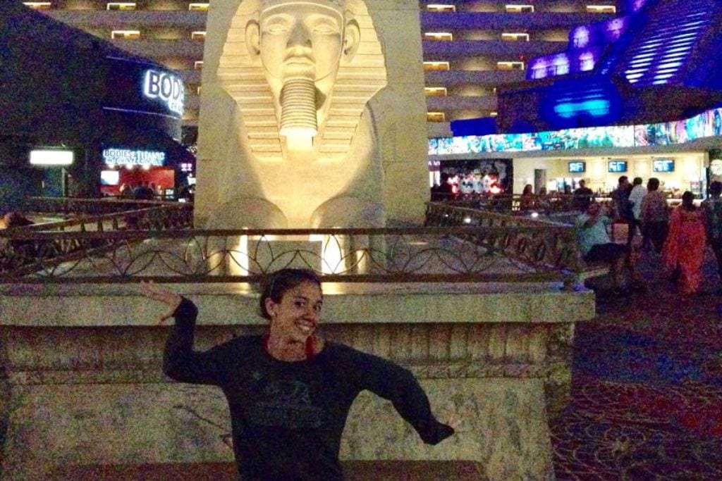 A smiling person poses with an outstretched arm in front of a large Sphinx replica inside the Luxor Hotel, Las Vegas, with a bustling atmosphere and dim lighting highlighting the Egyptian-themed decor. Luxor City in Egypt 2025 Guide to the Ancient City.