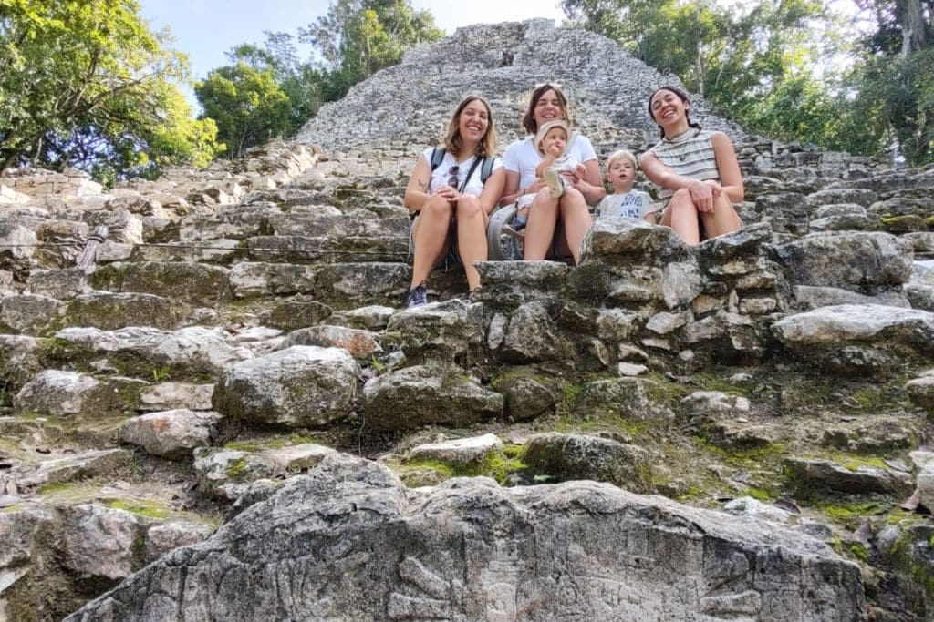 Your 2026 Spiritual Guide to The Pyramids of Teotihuacan Four smiling individuals seated on ancient stone steps of a Mayan ruin in Teotihuacan, with lush greenery in the background. Two adults and two children enjoy a sunny day out, creating a snapshot of family adventure at a historical site.