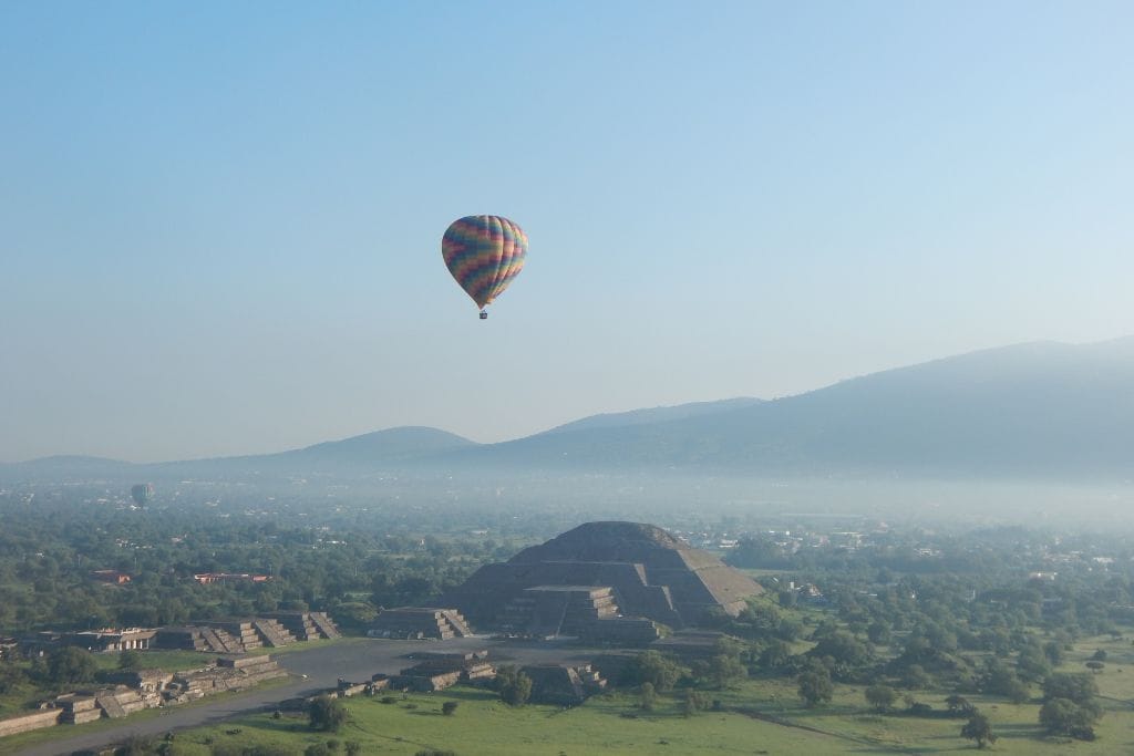 Your 2026 Spiritual Guide to The Pyramids of Teotihuacan A hot air balloon adorned with vibrant stripes floats in the clear sky above the lush landscape, with the Pyramid of the Moon at Teotihuacan visible in the mid-ground and a backdrop of rolling hills under a soft haze.