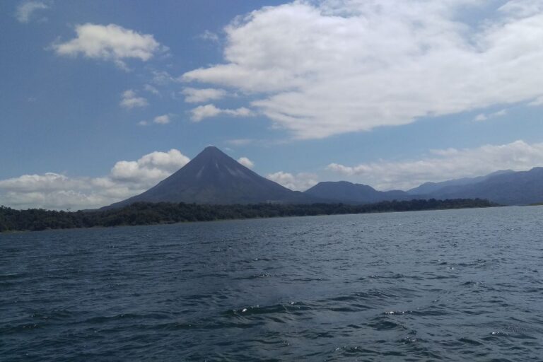 A serene view of the Arenal Volcano in Costa Rica, symbolizing tranquility and spirituality, with a calm lake in the foreground and a backdrop of lush hills under a blue sky with scattered clouds. 2025 Costa Rica Spirituality Guide of the Arenal Volcano Area!