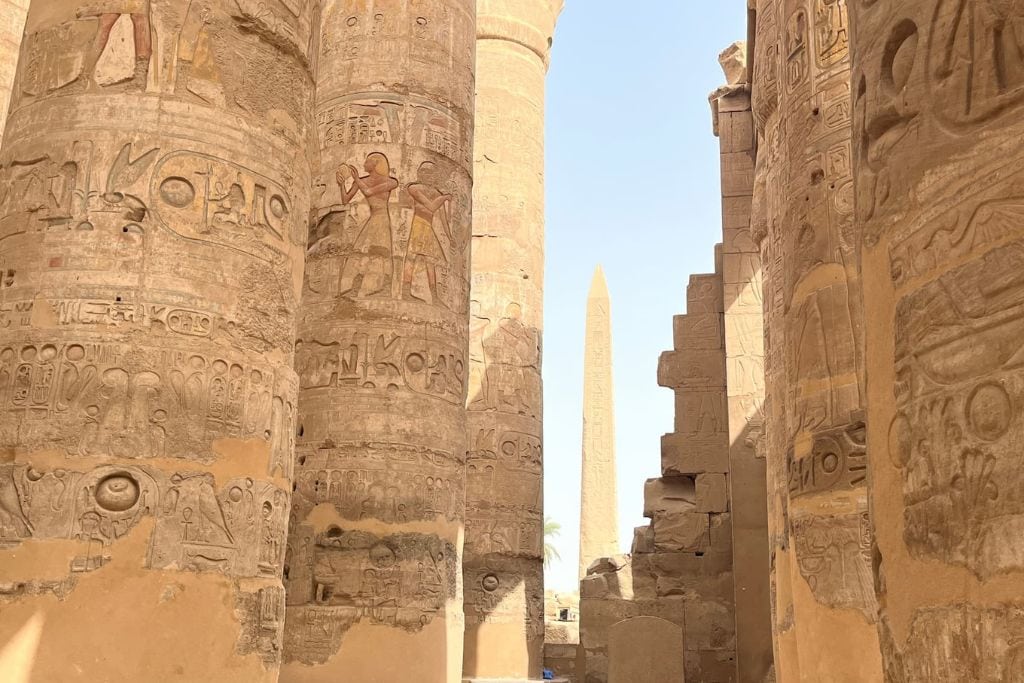 The towering columns of the Great Hypostyle Hall at Karnak Temple in Luxor are adorned with intricate hieroglyphs, with the silhouette of an ancient obelisk visible between the massive stone pillars against a sunlit sky. Luxor City in Egypt