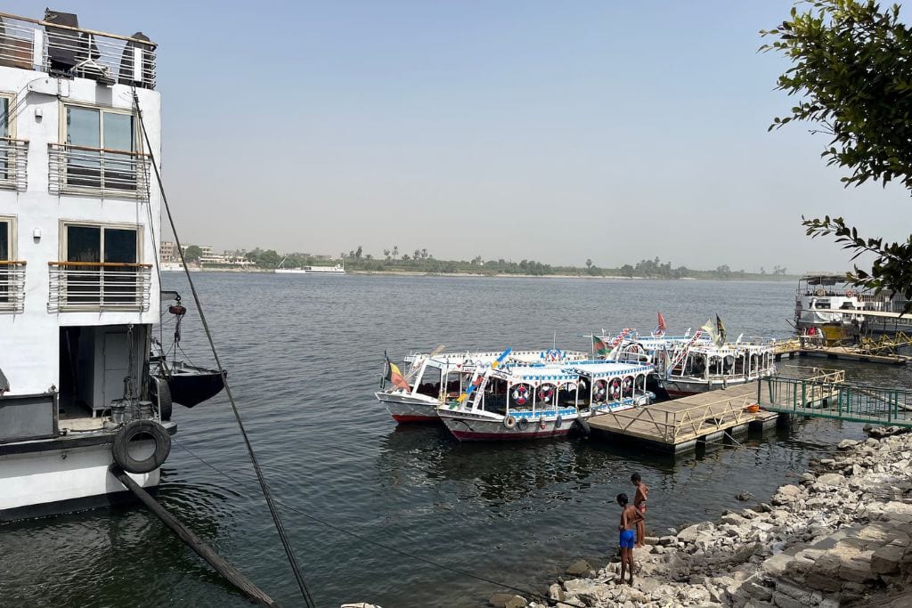 Riverbank of the Nile in Luxor, Egypt, with colorful, festively decorated boats moored at a pier, a child and adult standing by the water's edge, and a large white cruise ship anchored on the left.