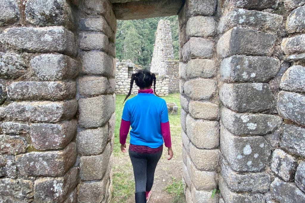 A hiker is seen from behind passing through a stone doorway on the Inca Trail, with a glimpse of Machu Picchu spiritual ahead. The person is dressed in a blue long-sleeve shirt, black leggings, and bright pink socks, contrasting with the grey stones and the greenery of the surrounding landscape.