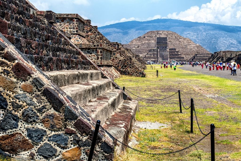 Your 2026 Spiritual Guide to The Pyramids of Teotihuacan The Pyramid of the Moon at Teotihuacan with tourists walking along the Avenue of the Dead, under a clear blue sky with distant mountains in the background.