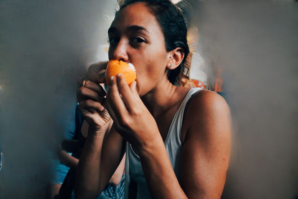 Your 2026 Spiritual Guide to The Pyramids of Teotihuacan A woman in a casual white tank top smelling a fresh orange with a slight smile, creating a sense of enjoyment and refreshment, with blurred figures in the background. The Pyramids of Teotihuacan.