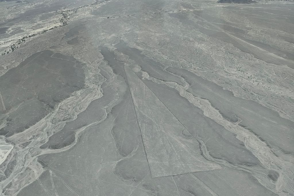 Why are the Lines of Nazca a Mystery of Peru? Expansive aerial view of the Nazca desert in Peru, displaying a vast network of the Nazca Lines with trapezoidal shapes and intricate geometric lines weaving through the arid landscape, under a hazy sky.