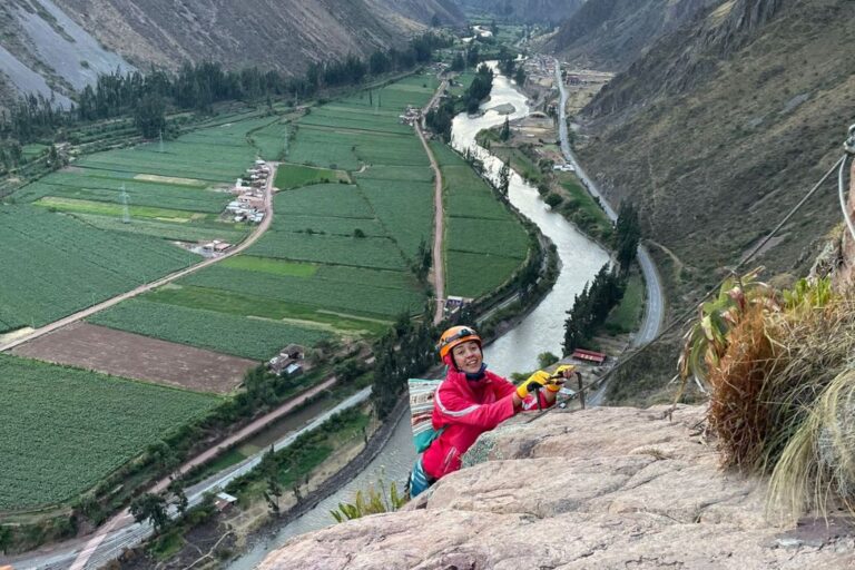 Adventurous climber in a bright pink jacket and orange helmet pauses to smile for the camera on a rocky outcrop, with the sweeping green fields and winding river of the Sacred Valley below, promoting Sacred Valley Spiritual Retreats.