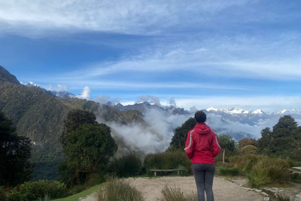 A person stands with their back to the camera, gazing at the breathtaking view of cloud-wreathed Andean peaks from the Machu Picchu. The sky is a clear blue above the rugged green landscape, and the individual is dressed for adventure with a red jacket and grey leggings, embodying the spirit of exploration.