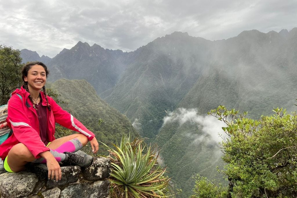 A joyful woman sits atop a stone wall along the Inca Trail, wearing a red rain jacket and bright pink socks, with a beaming smile. She's surrounded by the dense greenery of the Andes and low-hanging clouds, capturing the essence of the ancient trail's majestic and mystical atmosphere.