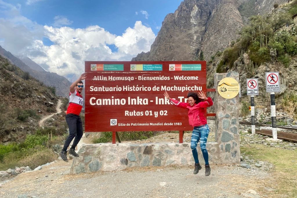 Two joyous travelers posing in front of the Camino Inka - Inka Trail sign at the historical sanctuary of Machupicchu in Peru. One is jumping with raised arms, the other waving, with the majestic Andean mountainscape in the background and clear prohibitory signs regarding the use of bridges and trails on the right.