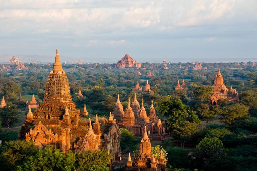 Is Bagan a Spiritual Place? Discover Why Should You Visit Bagan Myanmar in 2026! The image features a vibrant landscape of Bagan, Myanmar, showcasing numerous ancient Buddhist temples and stupas. These historic structures are bathed in the warm light of early morning, highlighting their intricate brickwork and pointed spires against a backdrop of lush greenery and a clear blue sky.