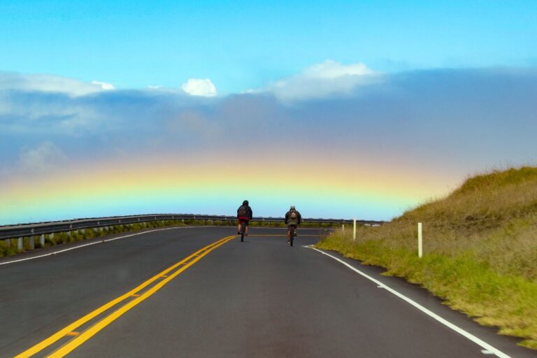 Two cyclists riding on the winding road of Haleakalā Volcano Hawaii with a vibrant rainbow arching across the sky in the background. The cyclists are in motion, and the scene captures a sense of adventure against the dramatic backdrop of a Hawaiian landscape.